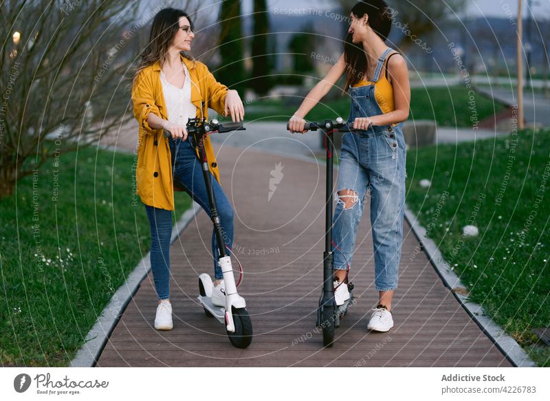 Crop smiling girlfriends with scooters interacting on urban footpath street style cheerful spend time weekend electric town lawn women trendy casual wear