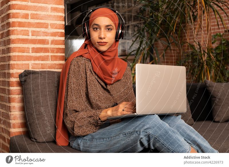 Muslim woman working on laptop on sofa gadget headphones using hijab surfing content watch couch computer internet browsing young muslim female netbook ethnic