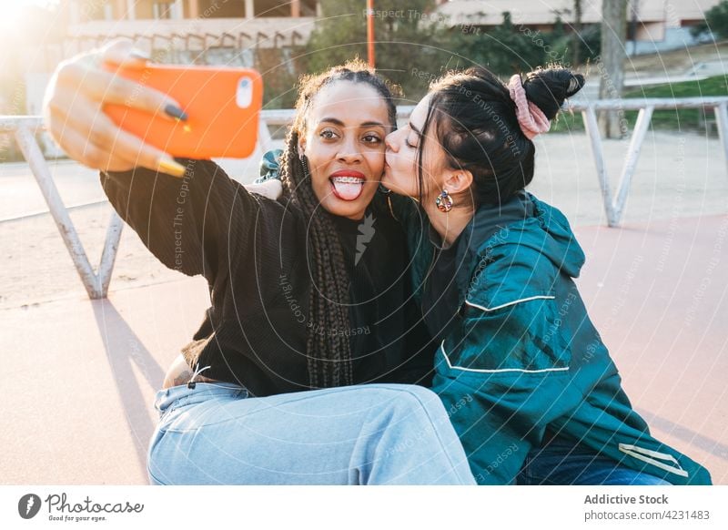 Multiracial lesbian couple kissing while taking selfie on smartphone outdoors same sex tongue out love memory women using gadget device street cellphone