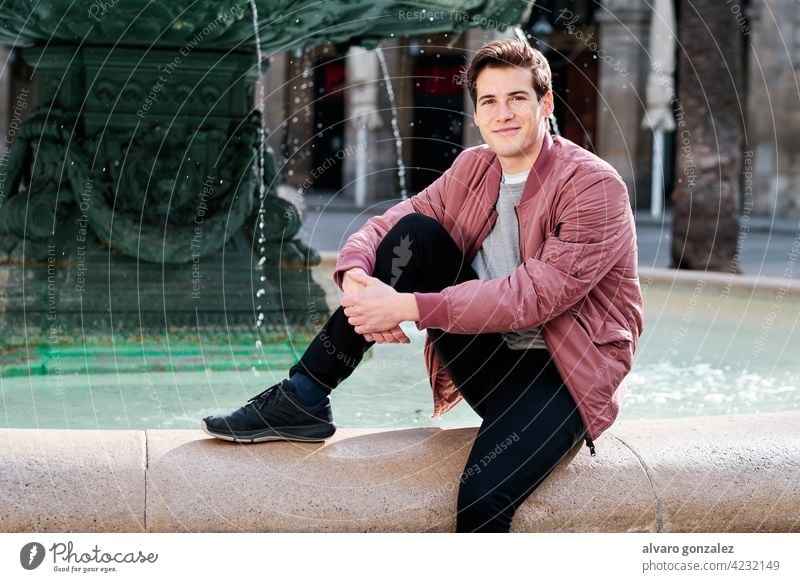 Young man sitting in a water fountain outdoors. portrait young urban city street casual style smile alone jacket clothes caucasian travel tourist outside