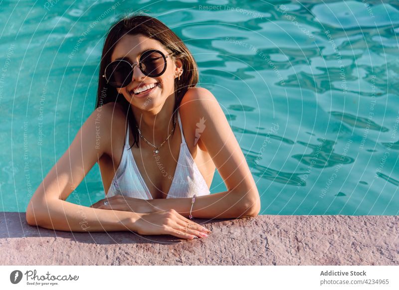 Smiling ethnic traveler in sunglasses in pool toothy smile woman accessory jewelry feminine vacation allure portrait gentle charming lean on hand cheerful