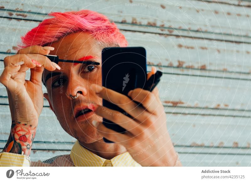 Stylish gay making up eyelashes against smartphone on gray background makeup mascara fashion unusual feminine portrait using man gadget device cellphone