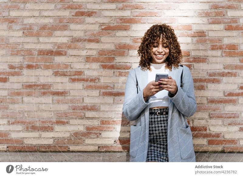 Cheerful ethnic woman on smartphone against brick wall toothy smile browsing cheerful communicate texting happy social media surfing message device positive