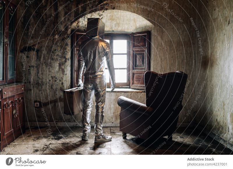 Faceless person in silver suit standing in shabby room suitcase concept chair relax incognito bizzare abandoned box window apartment comfort creative at home