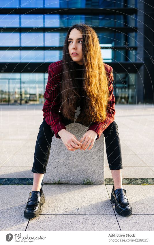 Stylish woman sitting against modern city building style trendy urban street confident unemotional appearance street style individuality serious young outfit