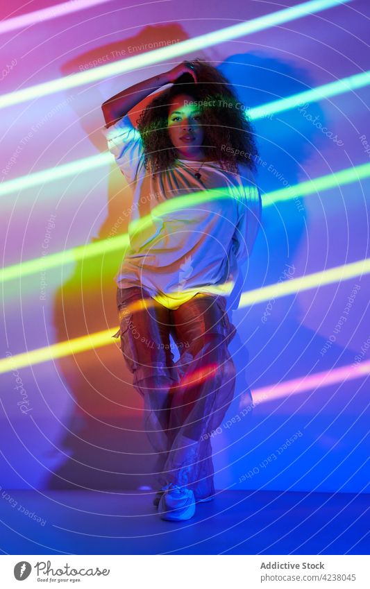 Trendy ethnic female dancer standing in neon lights woman style confident cool sunglasses energy posture informal lean young wall curly hair dynamic slim
