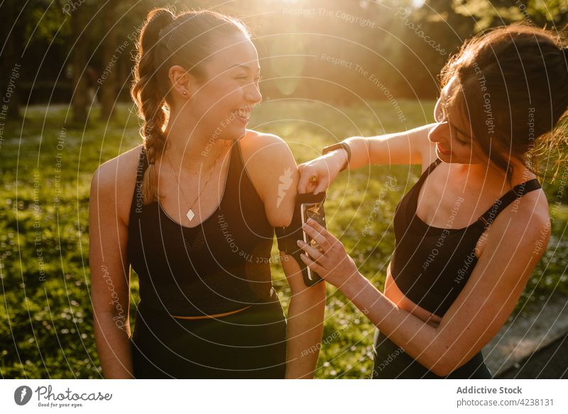 Smiling sportswoman touching smartphone armband on arm of girlfriend sportswomen put on help cheerful talk friendship sunshine sincere body fit park meadow