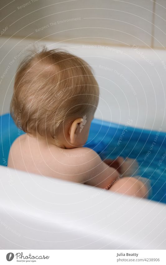 11 month old baby with back to camera sitting in a blue plastic bath tub;  blonde wispy hair - a Royalty Free Stock Photo from Photocase