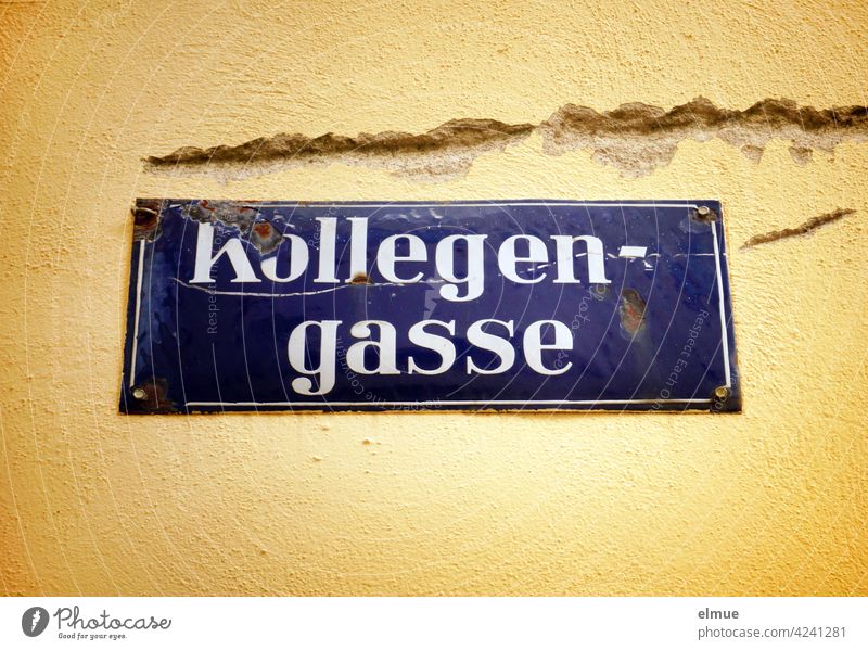 Damaged, blue enamelled street sign " Kollegengasse " at a likewise damaged yellow house wall / live street name corrupted Blue Yellow Street dwell Envy Anger