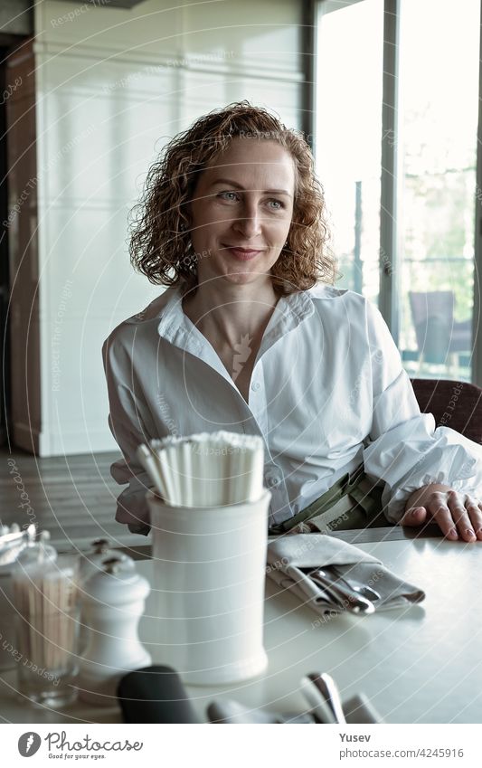 Beautiful smiling middle-aged woman in a white shirt in a restaurant. Portrait of an attractive curly haired lady in a cafe. Natural summer morning light. Front view. Vertical shot. Life style