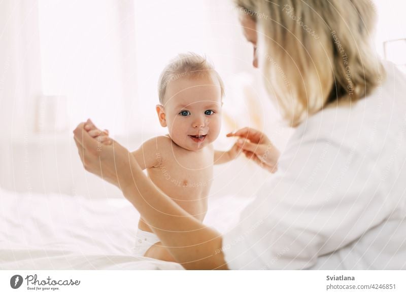 A cute baby is sitting on the bed with her arms outstretched and holding her mother's hands. Develop new skills, support and care child mom bedroom smiling