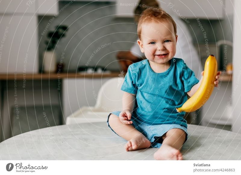 A little boy sits on the kitchen table with a banana in his hands and smiles small baby food healthy happy fresh toddler cute fruit nutrition vitamin delicious