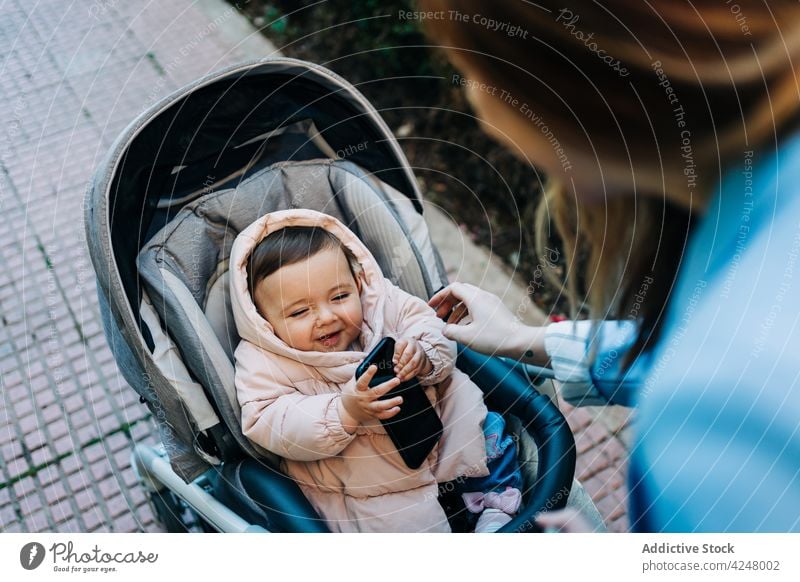 Adorable baby with smartphone sitting in stroller on street mother cute spring season curious lifestyle child funny warm clothes using carriage infant care