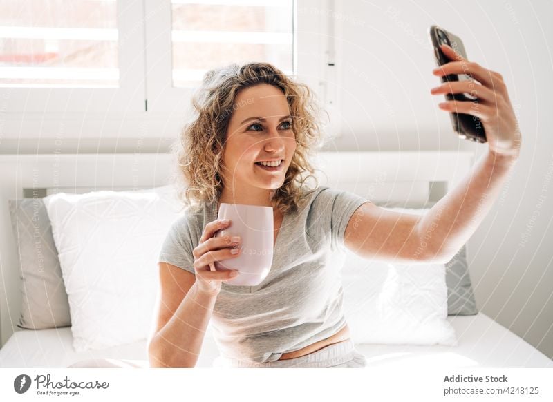 Smiling woman with cup taking selfie on bed cheerful toothy smile smartphone mug self portrait memory moment bedroom device mobile young cozy comfort using
