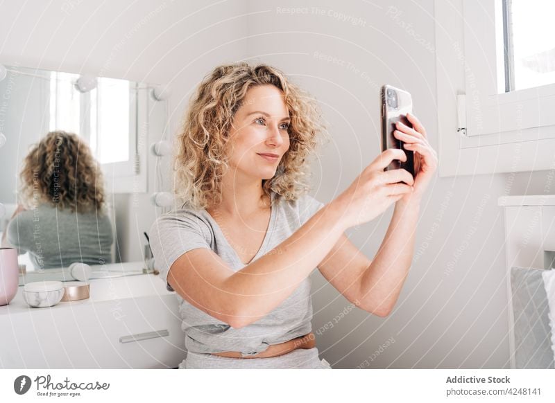 Smiling young woman talking selfie in bathroom smartphone smile positive social media take photo joy internet blogger digital mobile female curly hair blond