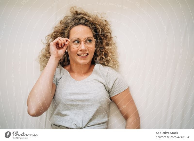 Delighted young woman adjusting eyeglasses while lying on bed smile happy comfort joy rest positive bedroom enjoy female curly hair blond casual delight relax