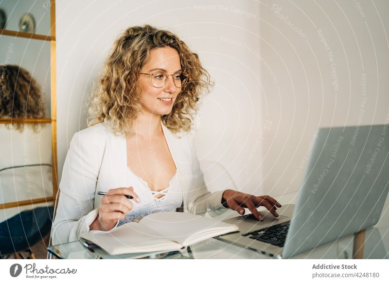Young woman writing in notebook and working on laptop at home take note freelance happy write smile remote online internet planner job using delight female