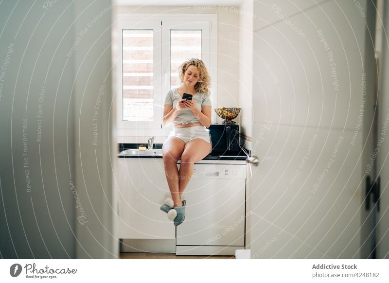 Cheerful woman using smartphone sitting on kitchen counter browsing content gadget happy modern smile young device positive style casual moment at home