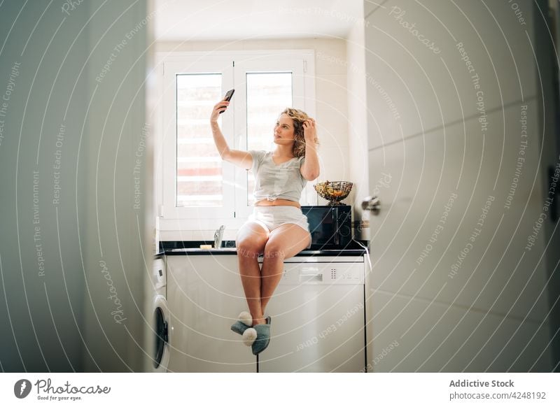 Cheerful woman taking selfie and sitting on kitchen unit touch hair smartphone self portrait using cheerful take photo content gadget modern young device