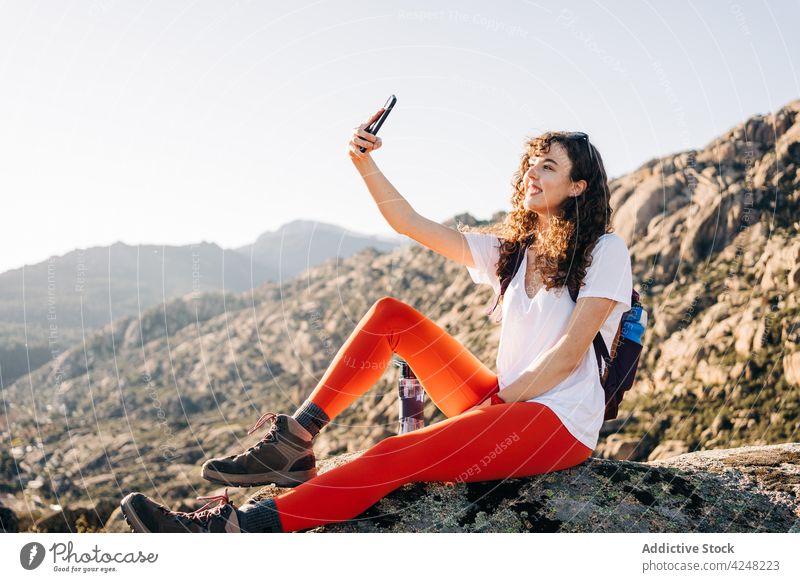 Cheerful young female backpacker taking selfie on mountain slope in sunlight woman smile traveler smartphone explore trekking take photo trendy nature content