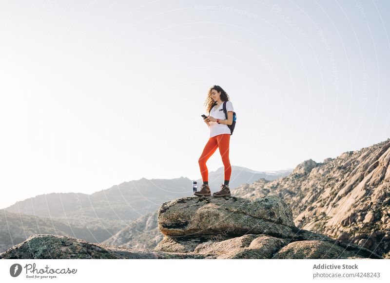 Cheerful young female backpacker using smartphone on mountain slope in sunlight woman smile traveler explore trekking trendy message browsing nature content