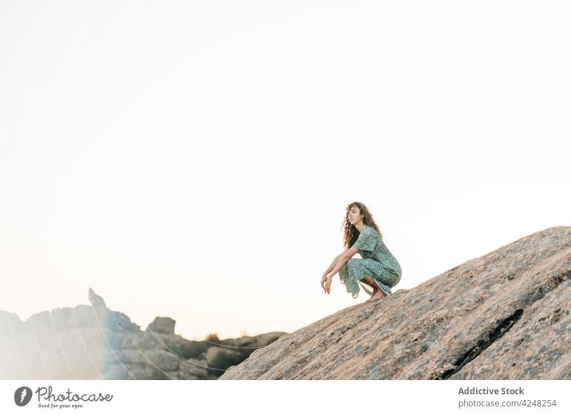 Smiling barefoot woman in sundress crouched on rocky hill cheerful highland nature toothy smile carefree attractive content positive young mountain summer stone