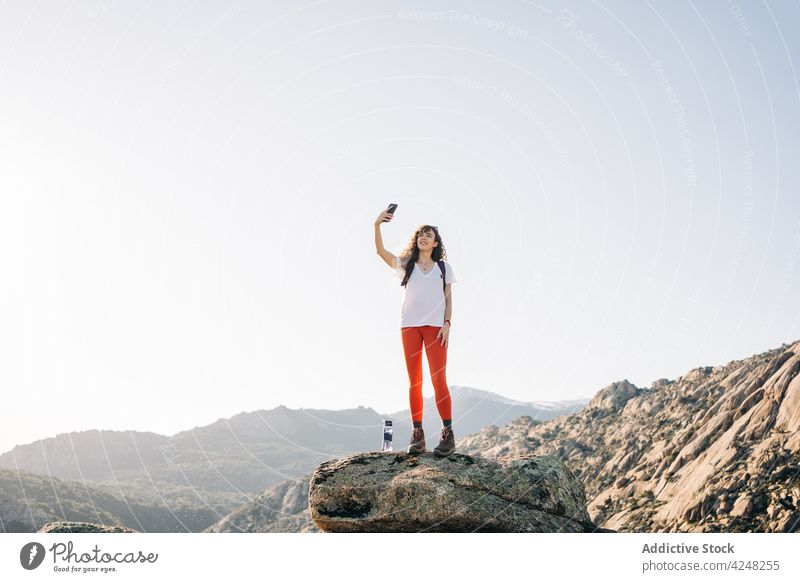 Cheerful young female backpacker taking selfie on mountain slope in sunlight woman smile traveler smartphone explore trekking take photo trendy nature content