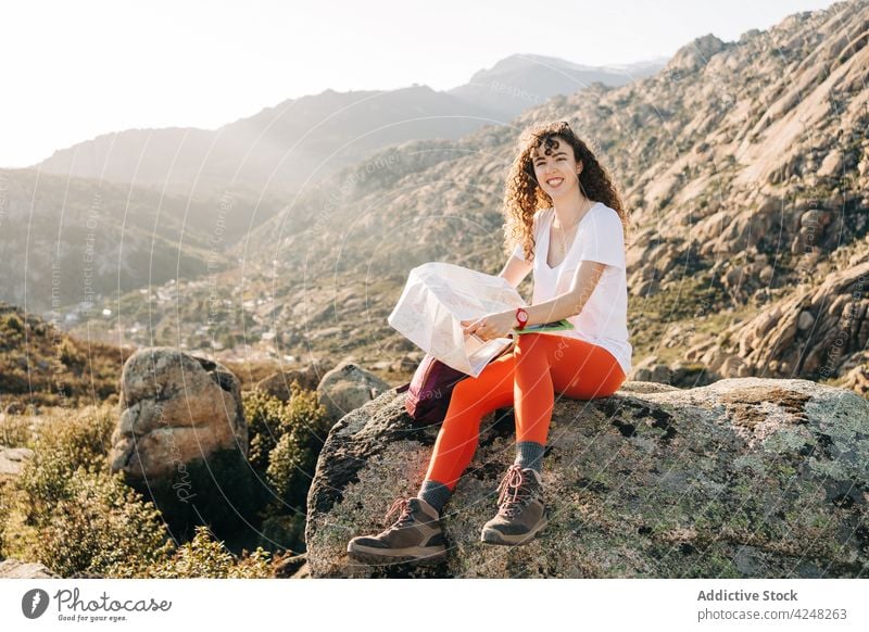 Joyful young woman sitting on mountain slope reading map hiker valley positive happy traveler joy vacation journey female curly hair enjoy nature smile cheerful