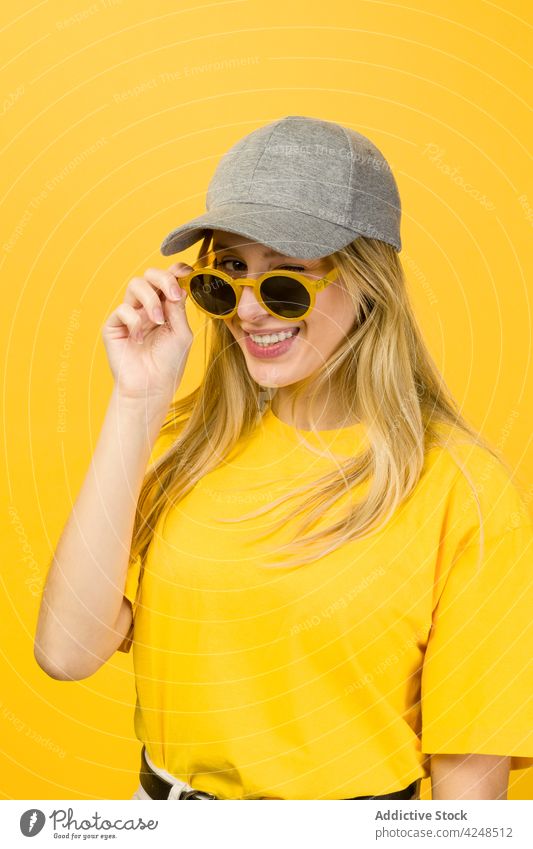 Woman with sunglasses in hands standing against yellow wall woman style vivid hipster street style cool appearance accessory sweater cap young funny bright