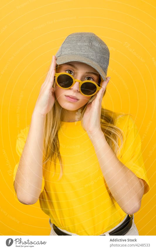 Woman with sunglasses in hands standing against yellow wall woman style vivid hipster street style cool appearance accessory sweater cap young funny bright