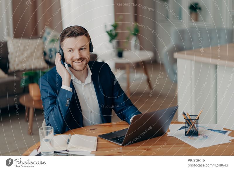 Smiling handsome young businessman in headset studying online at home work office talk call video computer suit education freelance laptop job casual lifestyle