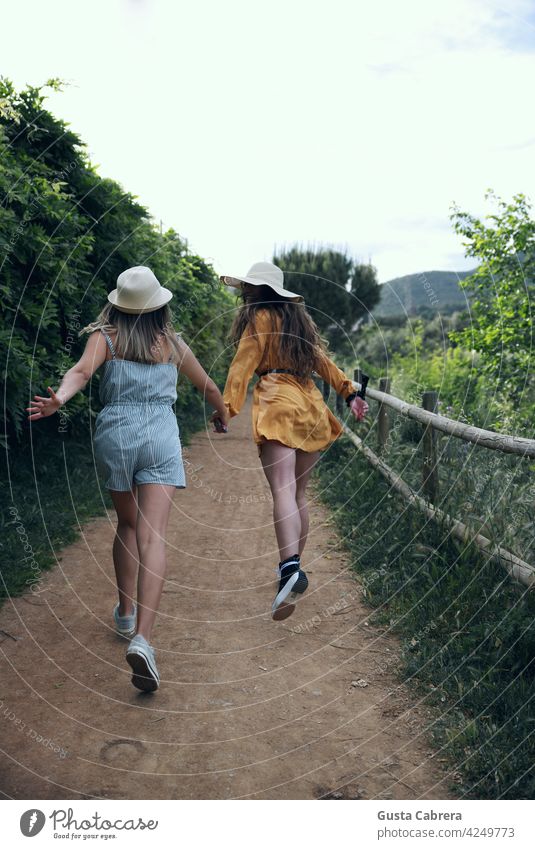 Two girls with hats smiling and running backwards along a path with many plants. happy talk young forest woman youth energy lifestyle park season outdoor