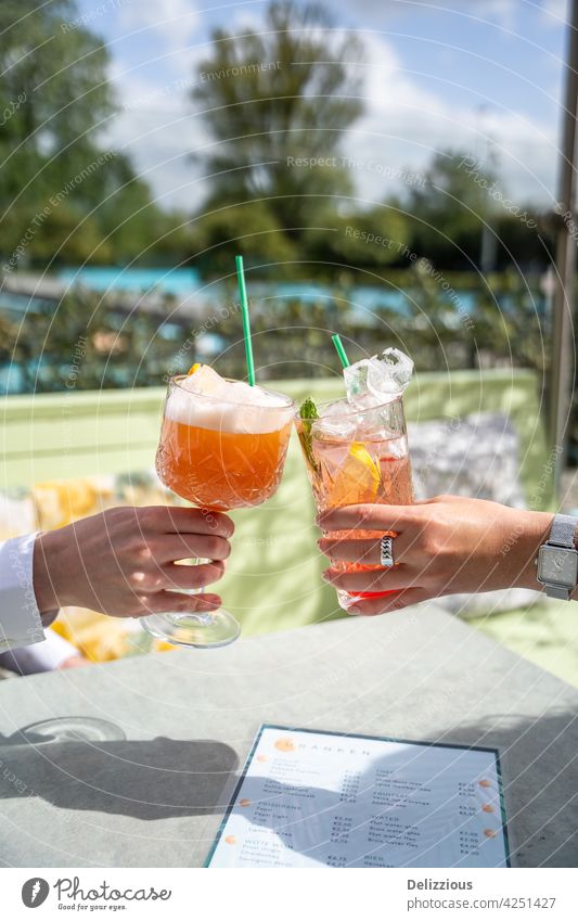 Two people cheering with pink cocktails outside on a terrace on a sunny day with swimming pool in the background cheers hands drink food and drink epicure bar
