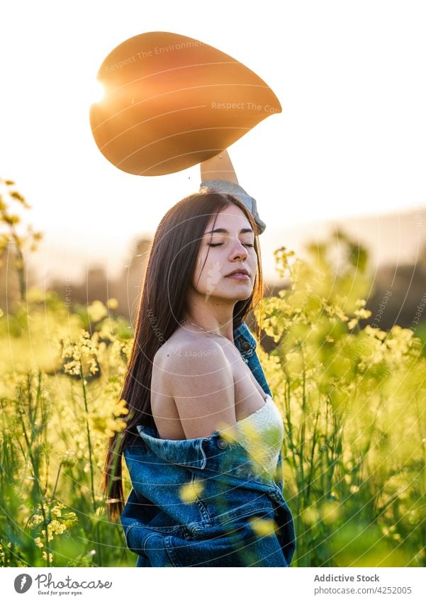 Woman raising arm with hat and standing on blossoming field woman sensitive arms raised eyes closed bare shoulders serene nature rapeseed feminine bloom