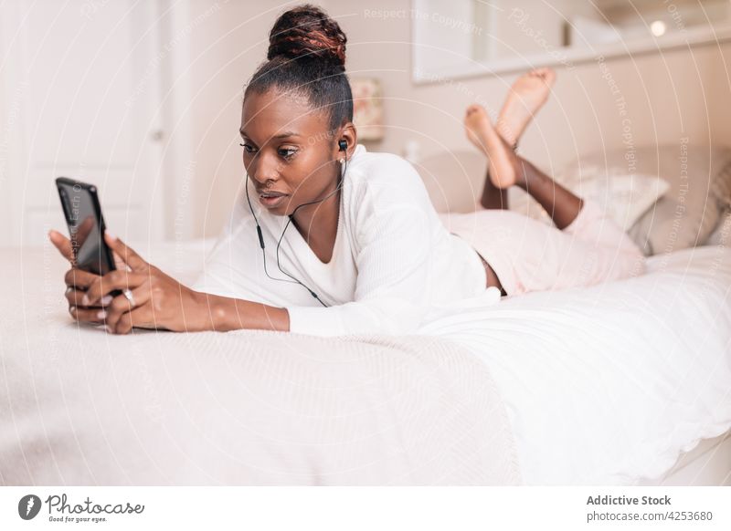 Positive young black woman using mobile and listening to music while lying on bed smartphone self assured smile relax enjoy recreation content female ethnic