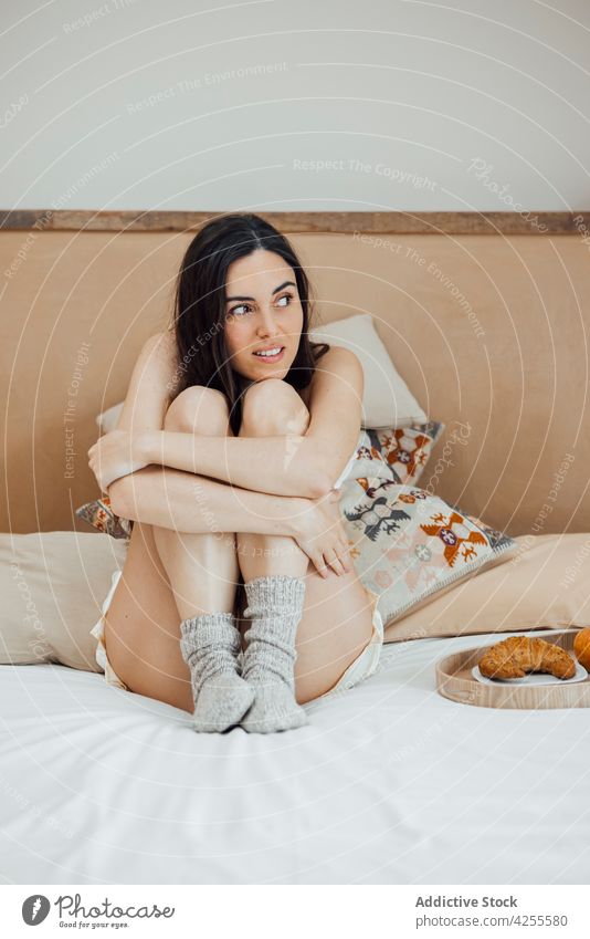 Smiling woman embracing knees and sitting on bed dreamy smile nightwear bedroom morning comfort serene lifestyle attractive brunette peaceful tender gentle