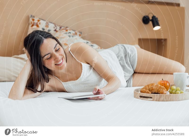 Woman with breakfast chilling on bed and using tablet woman morning relax tray calm browsing social media internet online home happy comfort gadget brunette