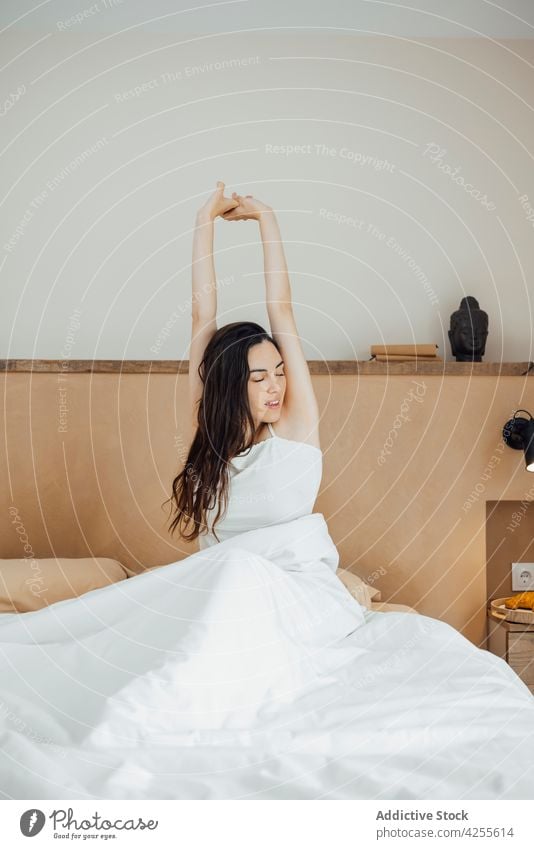 Smiling woman stretching arms after good sleep on bed wake up eyes closed toothy smile content awake bedroom morning glad comfort rest lifestyle cozy young