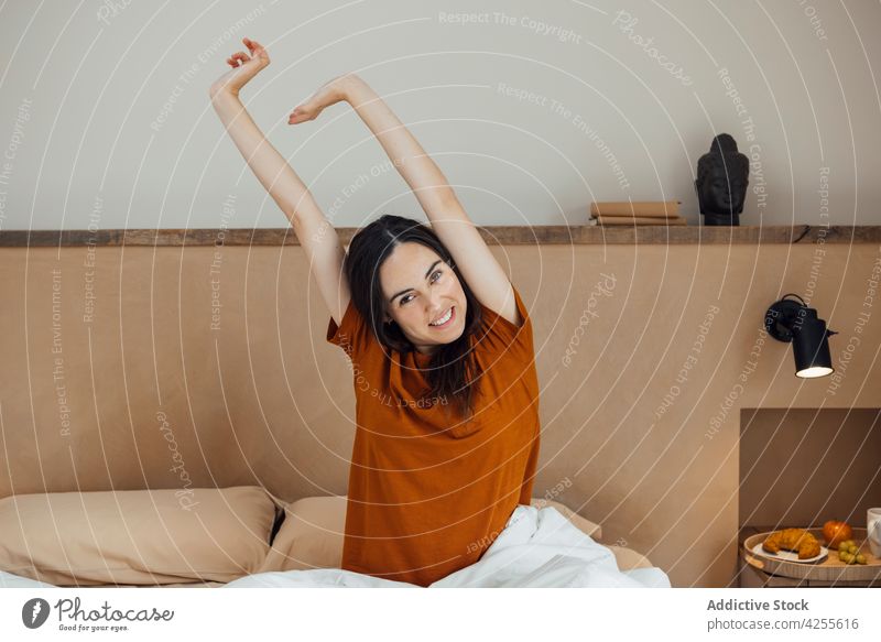 Smiling woman stretching arms after good sleep on bed wake up toothy smile content awake bedroom morning glad comfort rest lifestyle cozy young relax sit