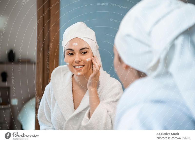 Woman in bathrobe applying cream on face in bathroom woman glad skin care mirror facial routine rejuvenate moisture touch face treat pamper cosmetology hygiene