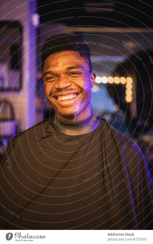 Cheerful black man sitting in barbershop client barber cape salon service positive masculine satisfied neon male customer optimist toothy smile expressive young