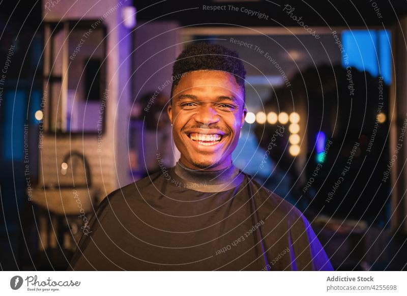 Cheerful black man sitting in barbershop client barber cape salon service positive masculine satisfied neon male customer optimist toothy smile expressive young