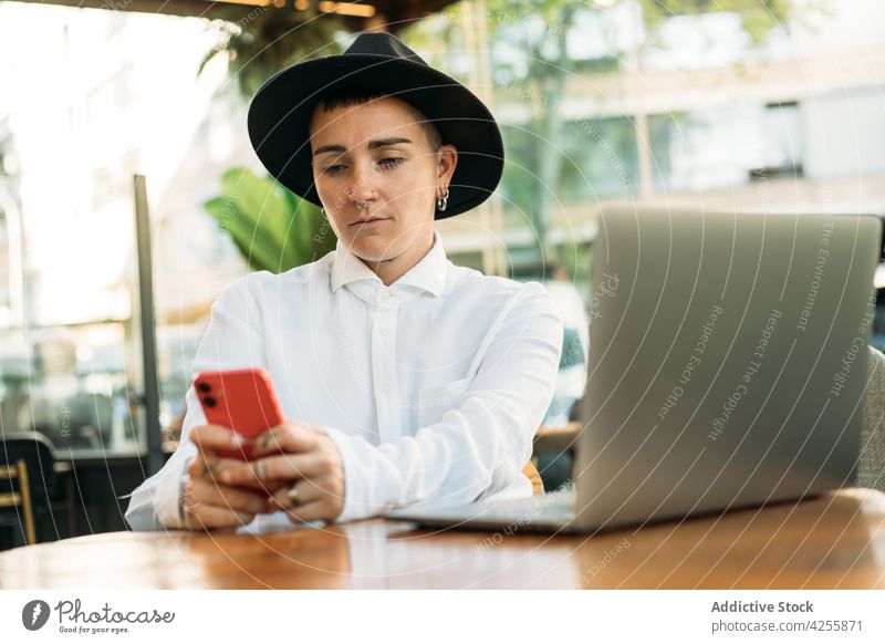 Tomboy browsing on cellphone while working on laptop woman tomboy using smartphone freelance transgender agender queer transsexual cafe netbook internet gadget