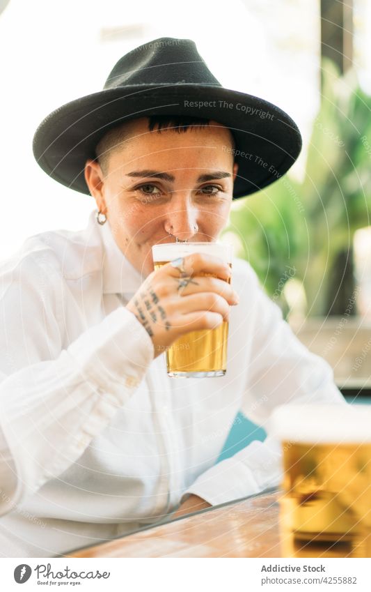 Smiling queer woman drinking beer at table tomboy booze taste alcohol glass brew transgender female agender identity foam beverage mug refreshment sit froth