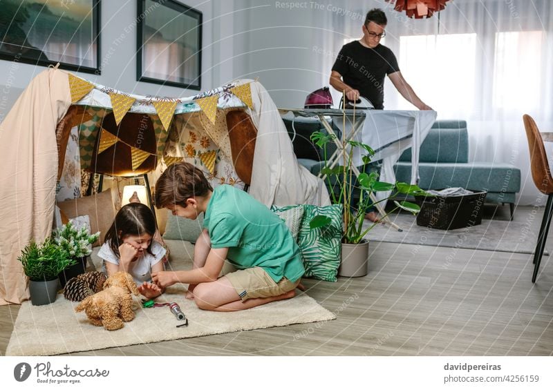 Father ironing while her children play camping at home father clothes tent family conciliation busy together living room househusband kid girl female people
