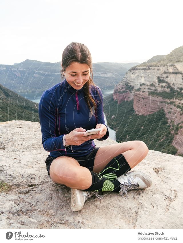 Cheerful young ethnic woman smiling and using smartphone on rocky cliff message smile canyon traveler trekking mountain enjoy rest legs crossed female hispanic
