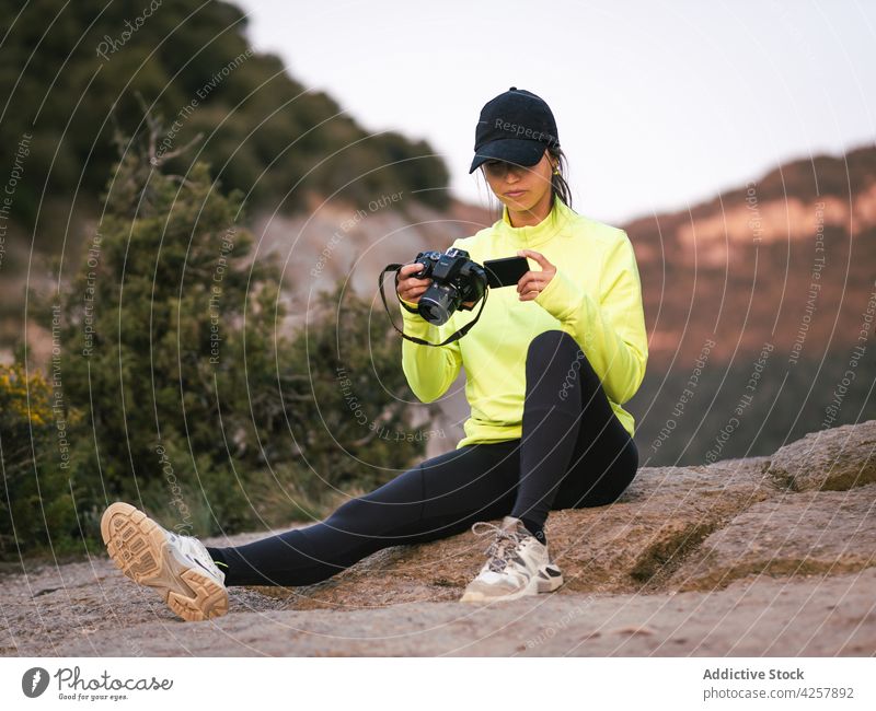 Young ethnic woman watching video while resting on stone during hiking trip traveler cliff nature trekking valley camcorder camera journey female young hispanic