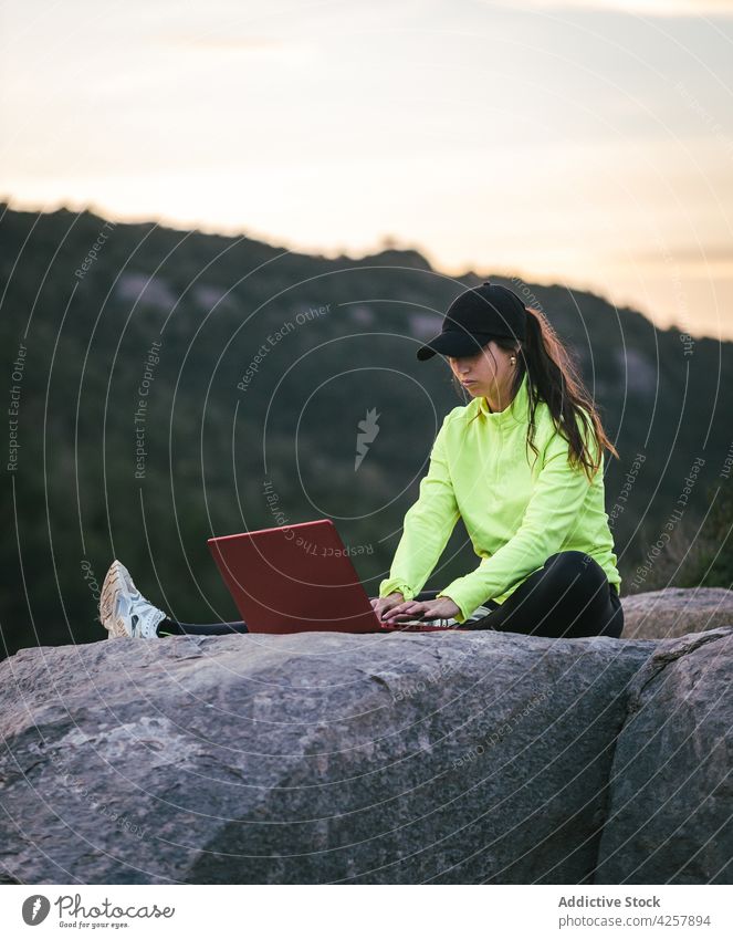 Female remote worker using laptop on rocky cliff above river at sundown woman mountain freelance browsing nature sunset chart travel female young casual cap