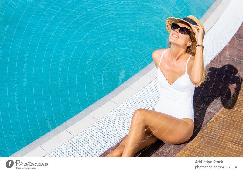 Happy woman sitting on poolside and sunbathing swimsuit happy hat resort chill vacation sunglasses swimwear rest paradise suntan enjoy holiday attractive slim