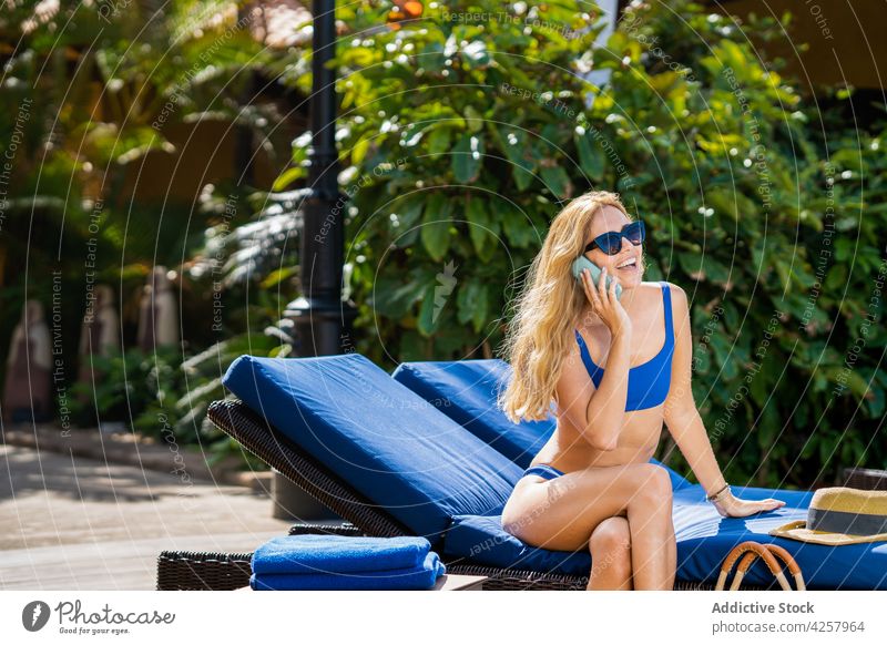 Joyful woman in bikini talking on smartphone on poolside deckchair phone call sunbath joyful toothy smile chat happy resort chill vacation sunglasses swimwear